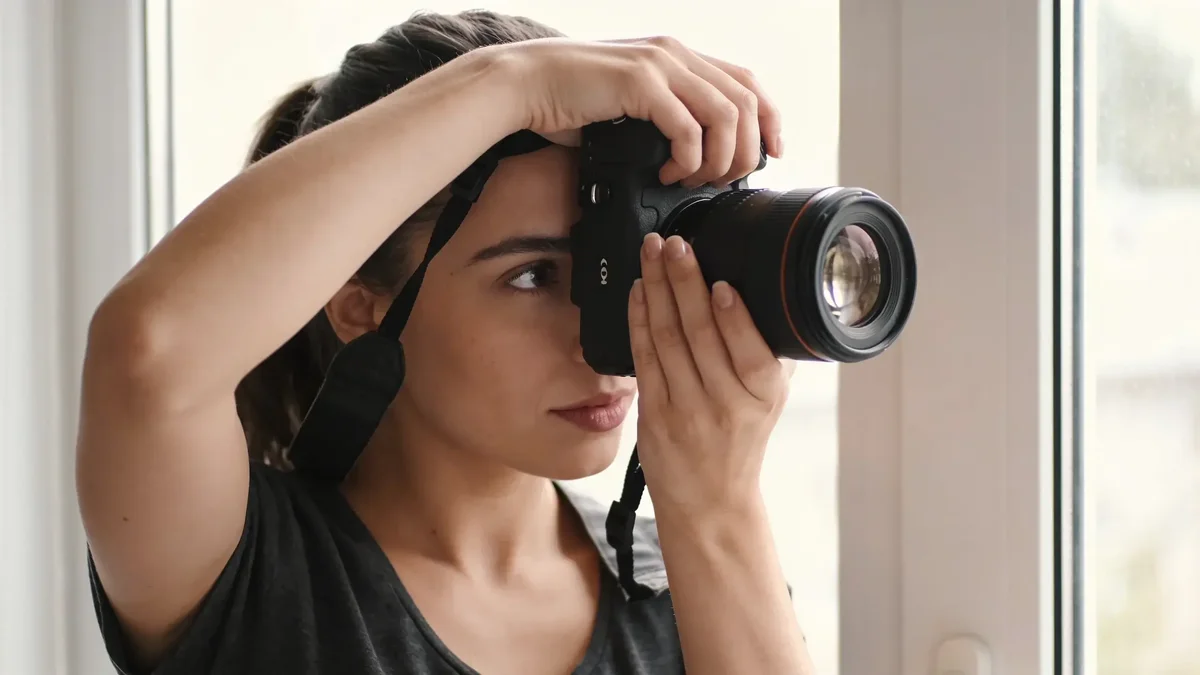 Fotografka pri okne mieri okom cez optický hľadáčik zrkadlovky, druhé oko má otvorené a sústredene komponuje záber
